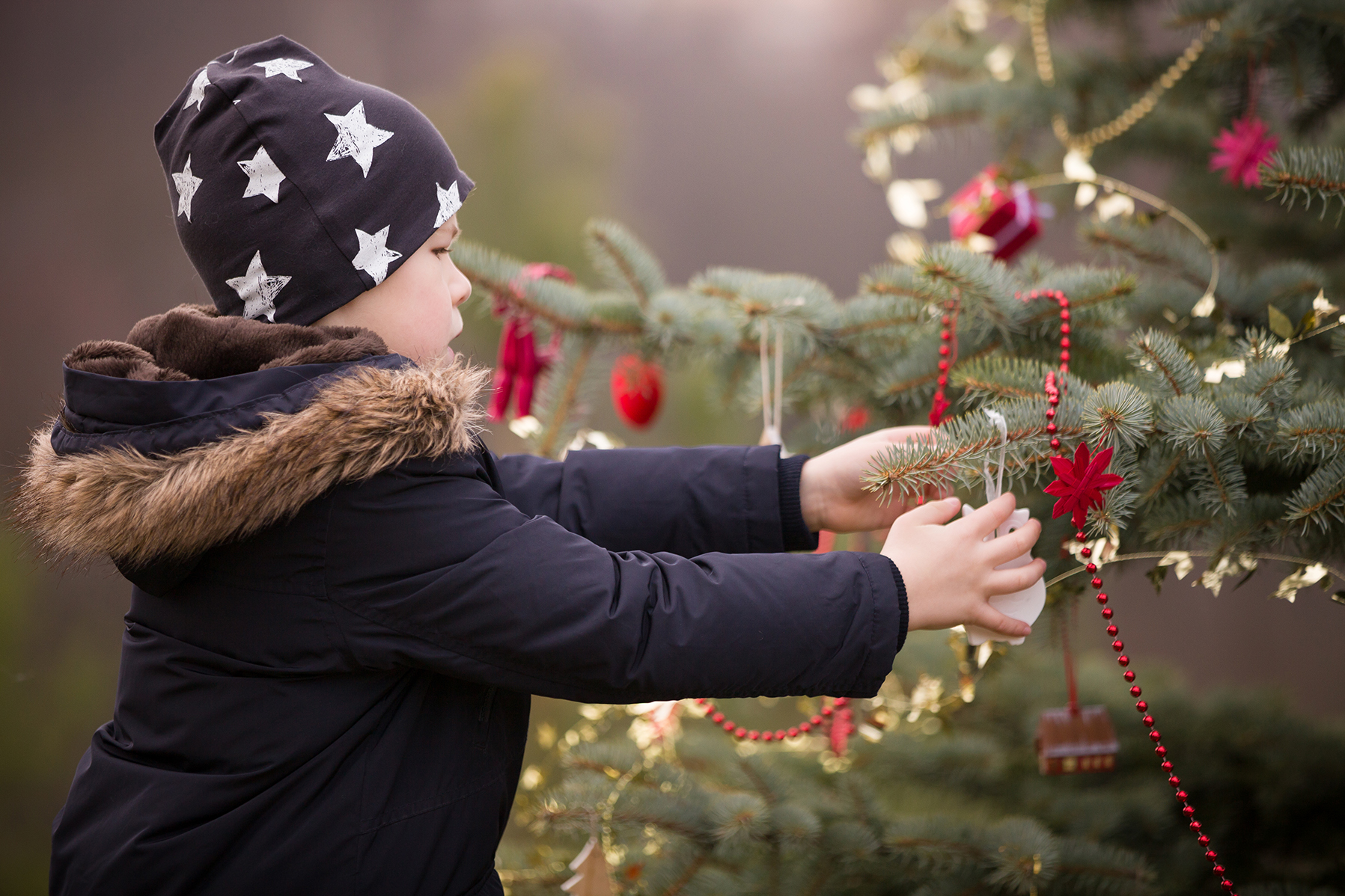 Portrait of cute kid boy in winter clothes decorating big Christmas tree outdoors. Child preparing for Christmas celebration.