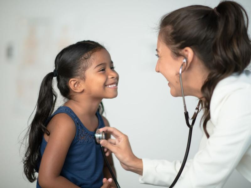 A beautiful little Hispanic girl sits in front of her doctor while at a routine check up. She has a big smile on her face and is dressed casually as she looks up at the doctor. The doctor is leaning in close and listening to her heart beat with her stethoscope.
