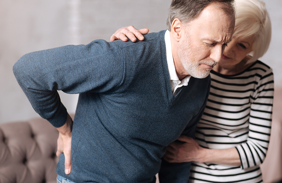 An older man holding his back and an older woman helping him