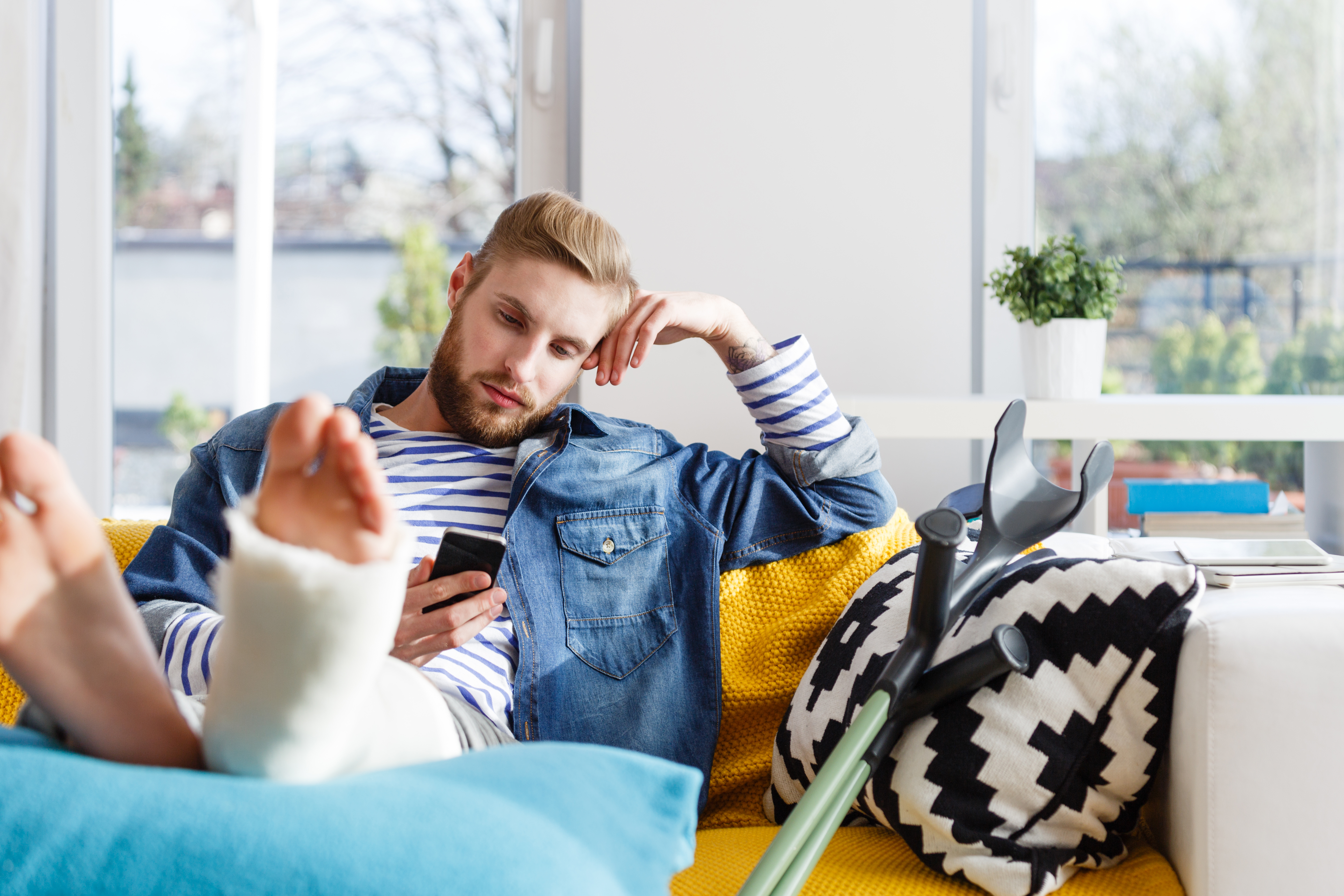 Young man sitting on the couch looking at their phone with their leg in a cast propped on a pillow