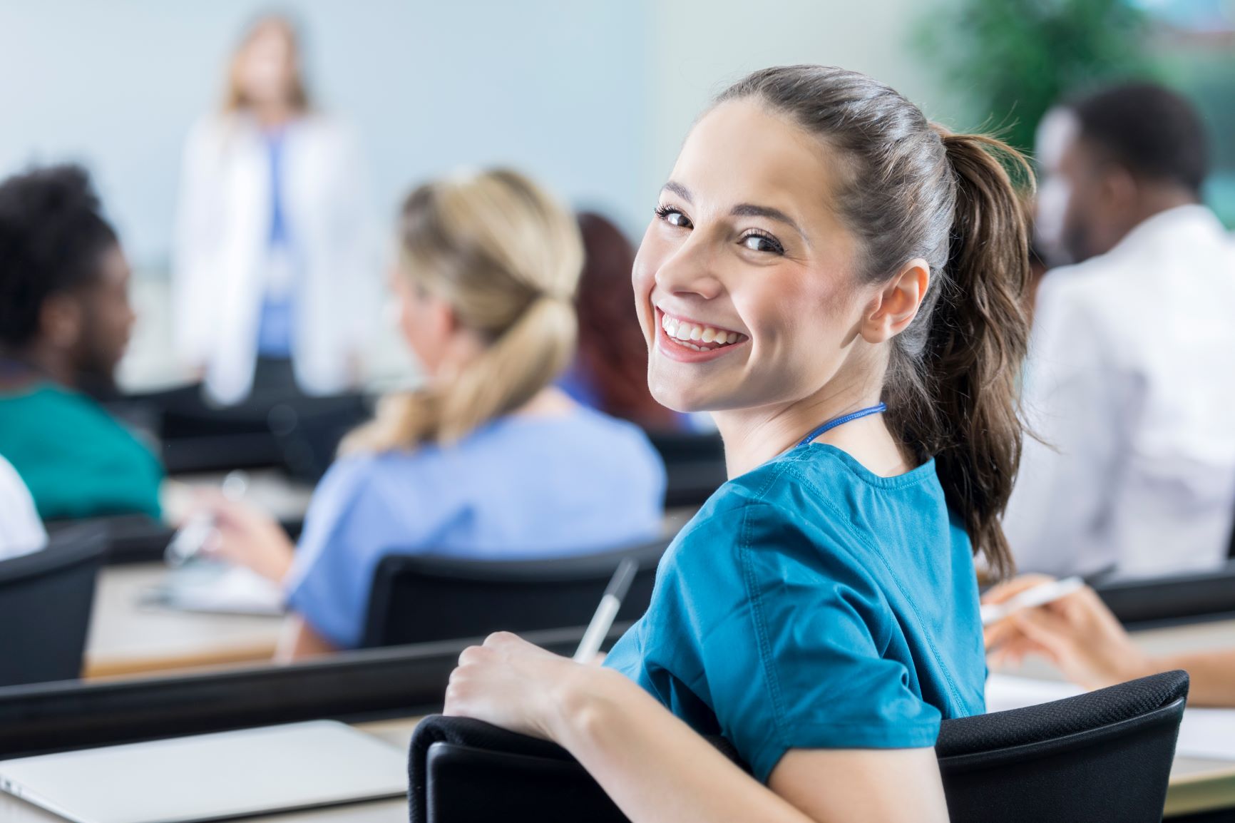 female medical student looking behind her should smiling while sitting at a desk in front of a female teacher in a lab coat