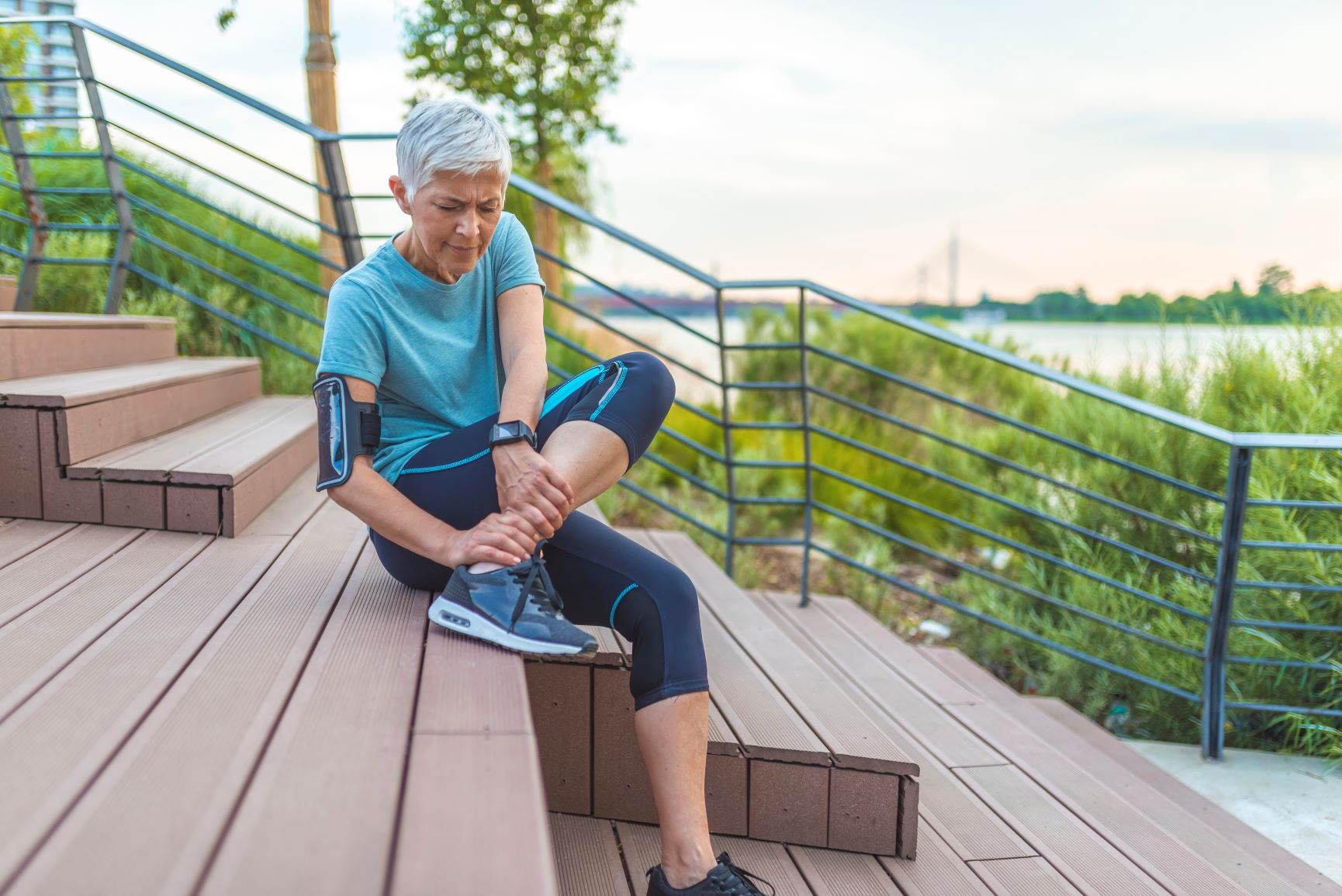 older woman in exercise clothes rubbing her ankle on steps outside