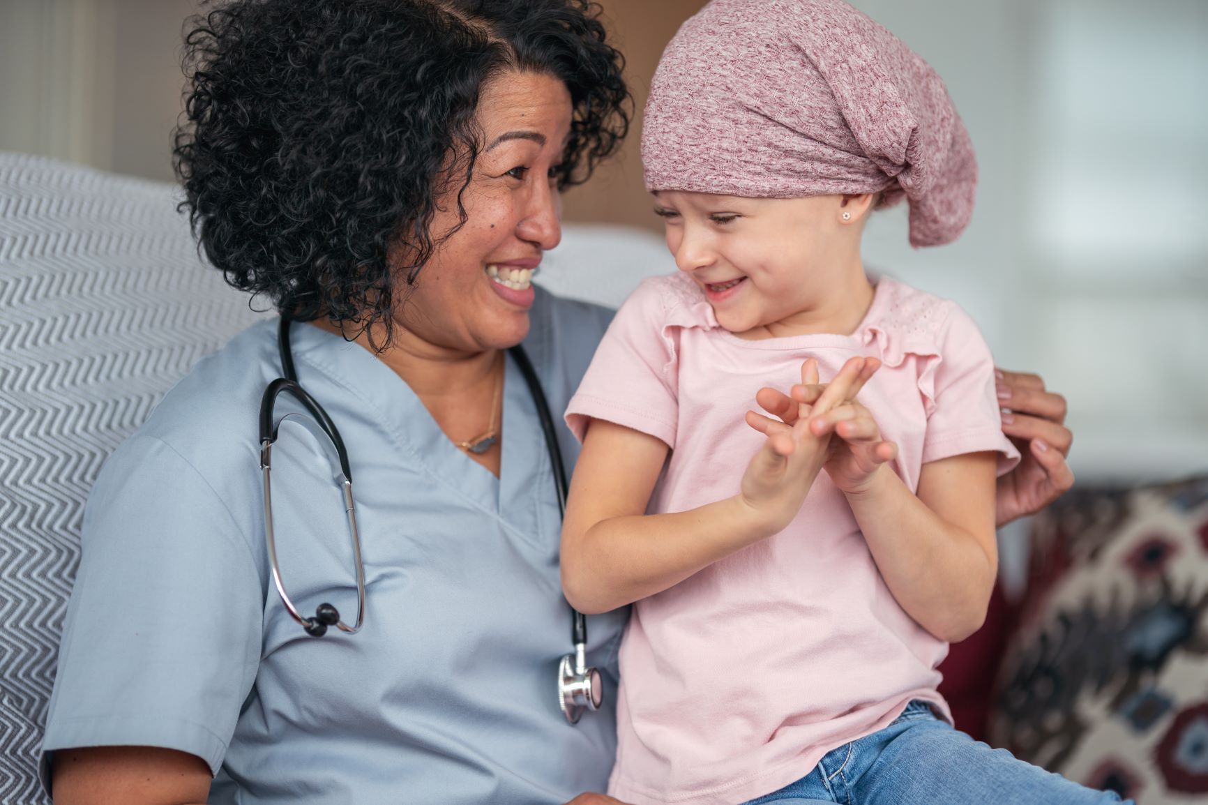 A female doctor of Asian descent sits with a child patient who is fighting cancer. The patient is a preschool age caucasian girl. She is wearing a pink bandana to hide her hair loss from chemotherapy treatment. The doctor is embracing the child. They are talking and the doctor is comforting the sick girl.