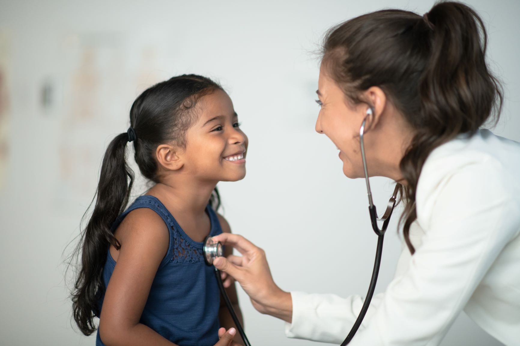 A beautiful little Hispanic girl sits in front of her doctor while at a routine check up. She has a big smile on her face and is dressed casually as she looks up at the doctor. The doctor is leaning in close and listening to her heart beat with her stethoscope.