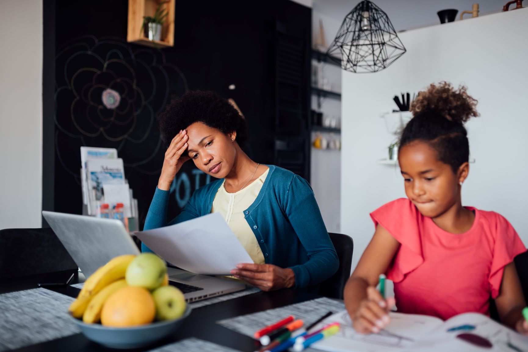 Woman looking stressed while working at the table next to a young girl who is coloring