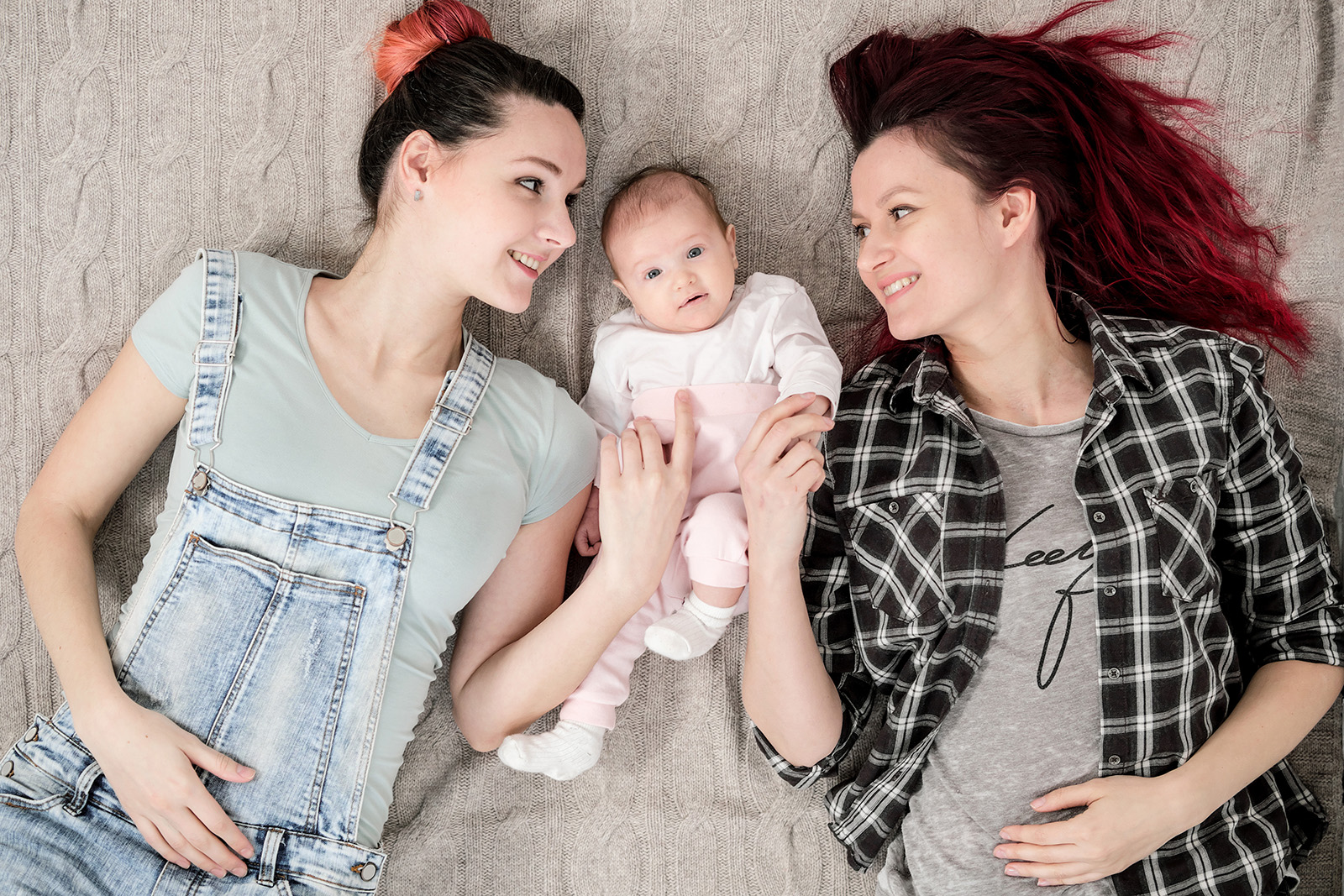 Two young women in casual clothes and with pink hair, a lesbian homosexual couple, lying on a rug with a child. Same-sex marriage, adoption.