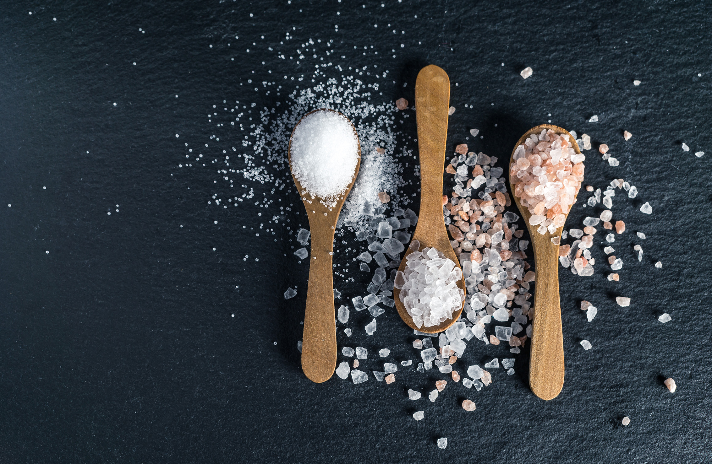Different types of salt. Sea, Himalayan and kitchen salt. Top view on three wooden spoons on black background