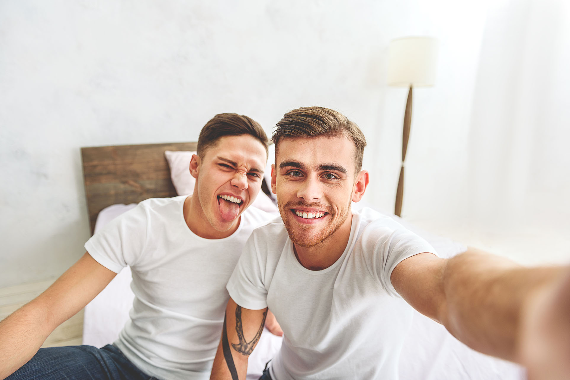two men smiling and sitting on a bed while taking a selfie.