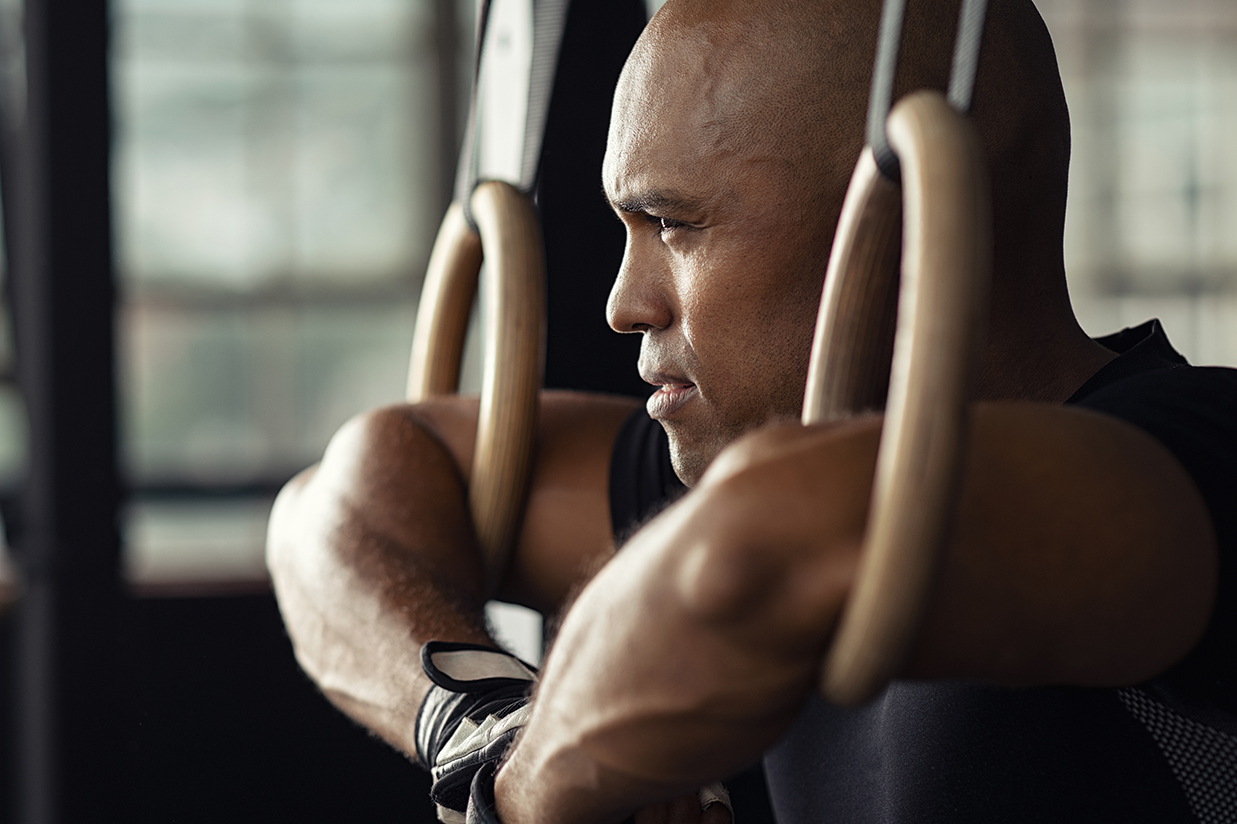 Strong athletic man resting on gymnastic rings. Black guy take a break at gym holding dip ring. Fit african man athlete training on gymnastic rings.