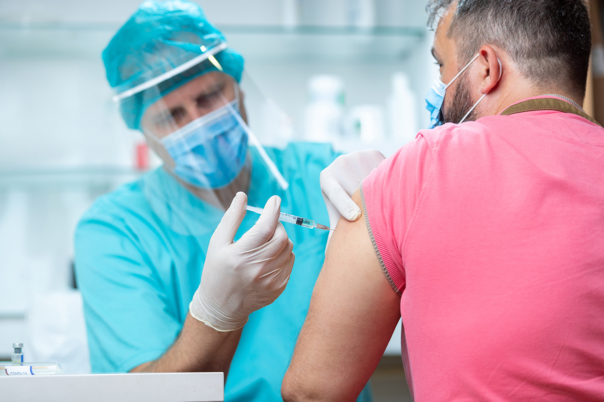 Doctor wearing protective visor and surgical gloves injecting COVID-19 vaccine into patient's arm