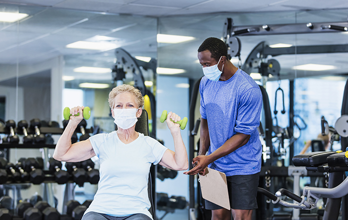 A senior woman in her 60s working out at the gym, lifting hand weights. Her personal trainer, an African-American man in his 30s, is watching her, adjusting her arm position.. She is exercising during the covid-19 pandemic. They are wearing protective face masks to prevent the spread of coronavirus.