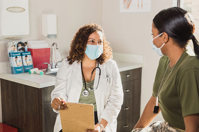 During an appointment in the midst of the coronavirus epidemic, the mid adult female doctor and the the female army soldier smile at each other through their protective masks.