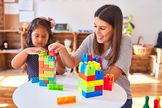 Beautiful teacher and toddler girl playing with construction blocks bulding tower at kindergarten