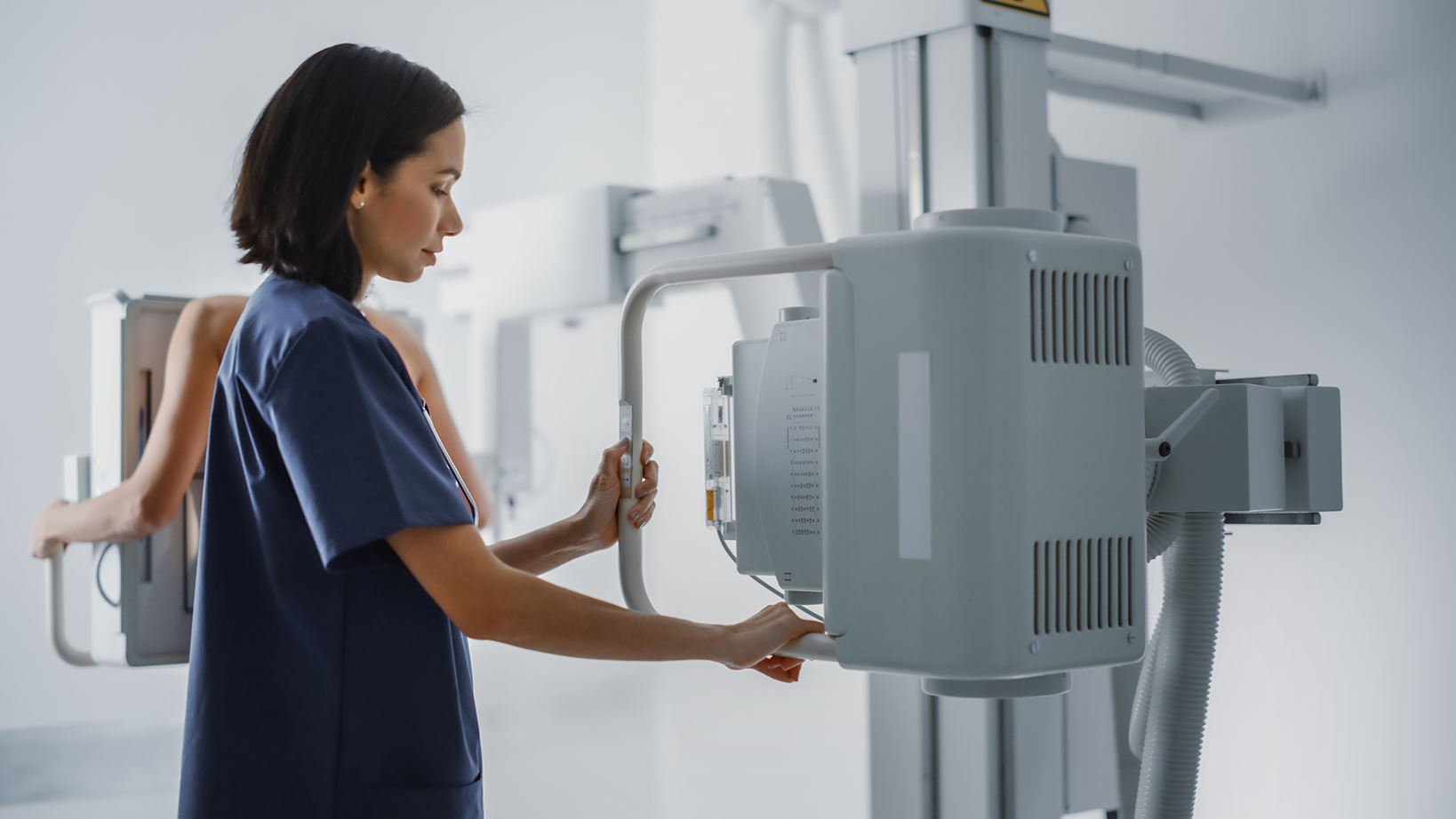 Hospital Radiology Room: Beautiful Multiethnic Woman Standing Topless Next to X-Ray Machine while Female Latin Nurse Adjusts it. Healthy Patient Undergoes Medical Exam Scanning with the Doctor's Help.
