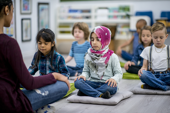 A multi-ethnic group of young school children are indoors in their classroom. They are sitting on pillows and doing yoga together. They are sitting with their hands in their lap.