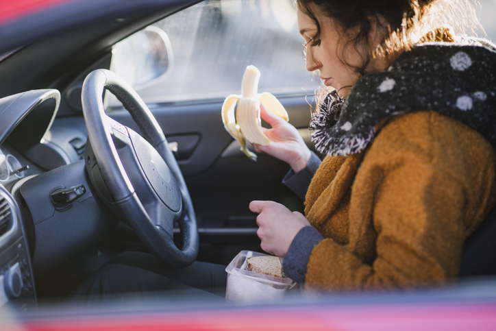Young woman is sitting in the driver's seat of her car eating a banana.