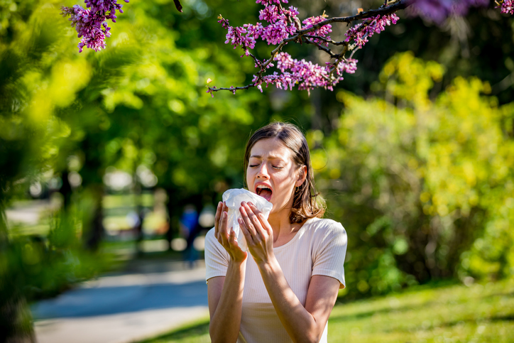 Young pretty woman blowing nose in front of blooming tree. Spring allergy