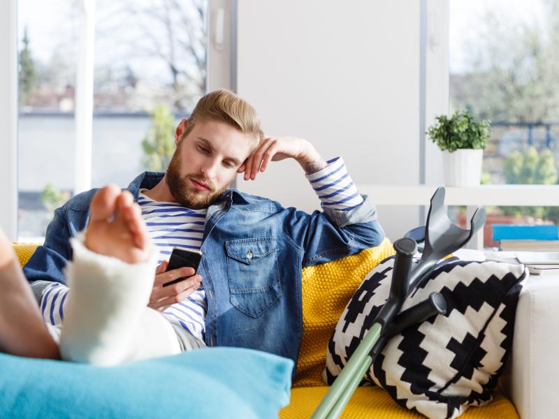 Young man sitting on the couch looking at their phone with their leg in a cast propped on a pillow