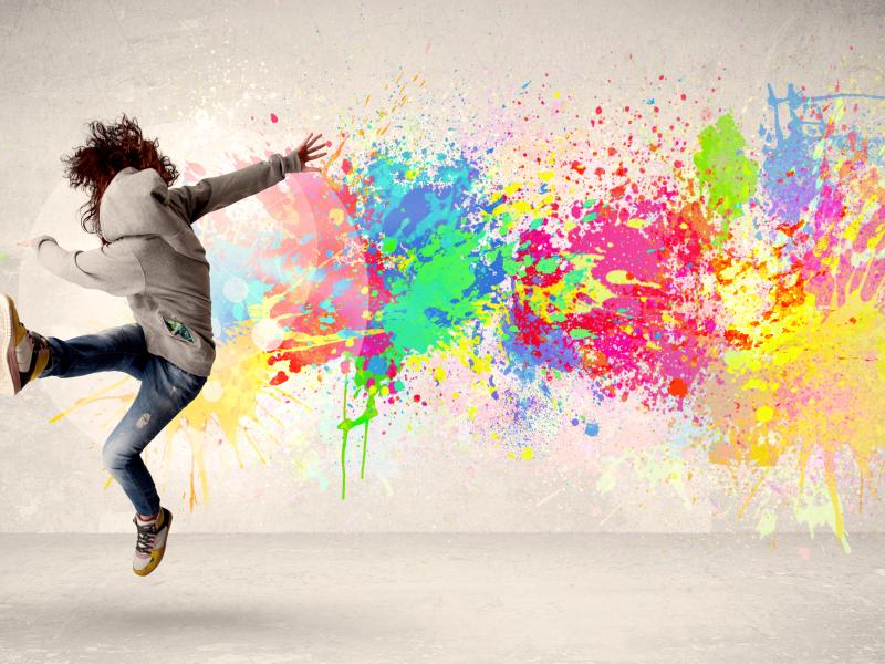 A person kicking one foot in the air with a colorful paint splatter on the wall behind them