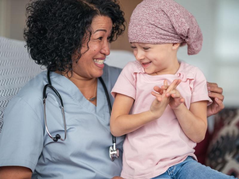 A female doctor of Asian descent sits with a child patient who is fighting cancer. The patient is a preschool age caucasian girl. She is wearing a pink bandana to hide her hair loss from chemotherapy treatment. The doctor is embracing the child. They are talking and the doctor is comforting the sick girl.