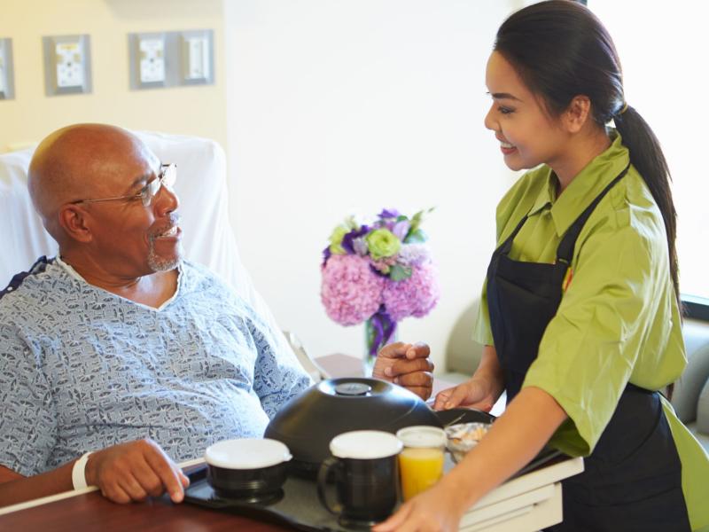 Senior Male Patient Being Served Meal On Tray In Hospital Bed