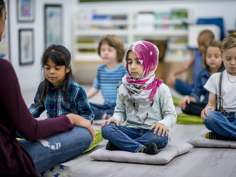 A multi-ethnic group of young school children are indoors in their classroom. They are sitting on pillows and doing yoga together. They are sitting with their hands in their lap.
