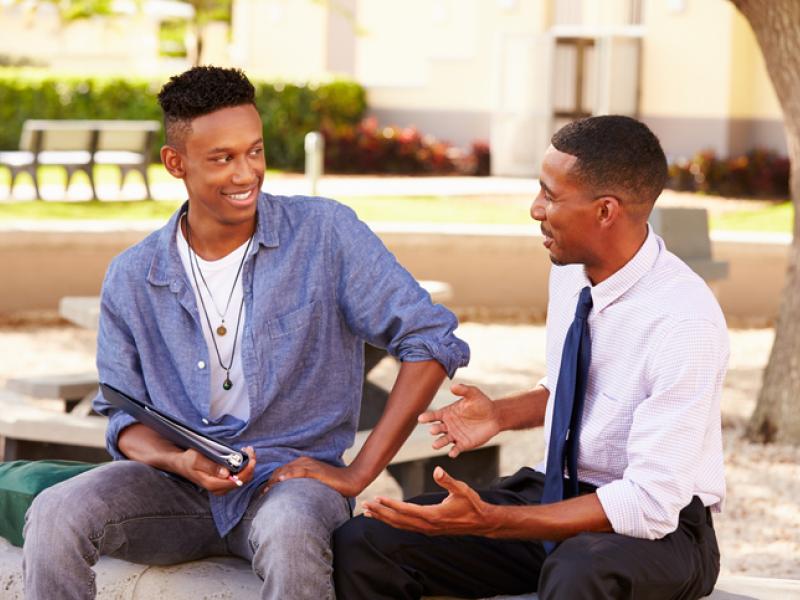 Teacher Sitting Outdoors Helping Male Student With Work Smiling