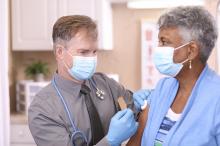 Mature adult doctor or healthcare worker, consultation with senior adult, African American female patient in office, hospital, or clinic setting. He gives patient vaccine or medicine injection. Both wear protective face masks. Coronavirus, medical exam, consultation.
