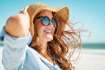 Side view of beautiful mature woman wearing sunglasses enjoying at beach. Young smiling woman on vacation looking away while enjoying sea breeze wearing straw hat. Closeup portrait of attractive girl relaxing at sea.