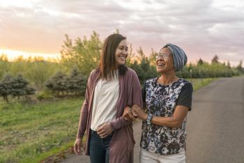A senior woman with cancer walks with her adult daughter at sunset down a rural road. 